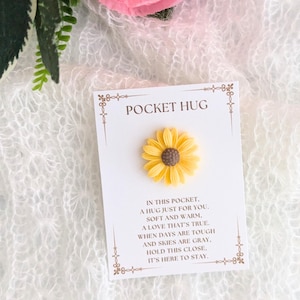 Peut inclure: Une petite fleur de tournesol jaune avec un centre brun est attachée à une carte blanche avec le texte "Pocket Hug" et un message sur un câlin juste pour vous. La carte est sur un fond texturé blanc et rose.