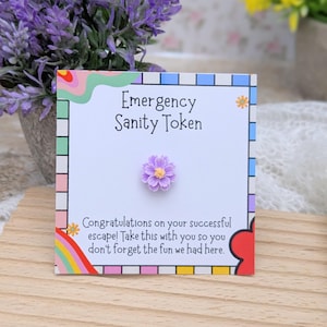 May include: A white card with the text "Emergency Sanity Token" and a small purple flower. The card also has the text "Congratulations on your successful escape! Take this with you so you don't forget the fun we had here."