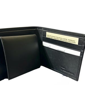 May include: Black leather wallet, open to display multiple compartments. The wallet has a billfold, card slots, and a coin pocket. A small tag with text is visible inside the wallet. The brand name is embossed on the bottom right.