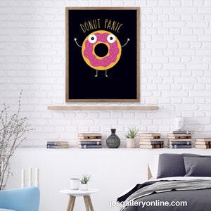 May include: A framed print with a black background and a pink donut with yellow eyes and arms. The donut has white sprinkles and the text "Donut Panic" in yellow letters.