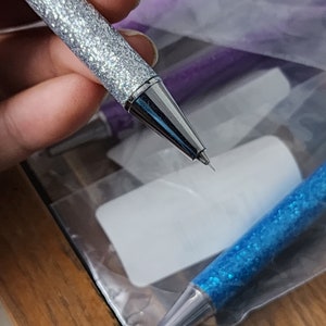 May include: A silver glitter mechanical pencil with a sharp point. The pencil is held in a hand and the tip is in focus.
