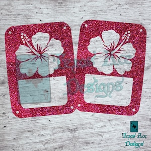 May include: Two pink glitter car license plate frames with a white hibiscus flower cutout. The frames are rectangular and have rounded corners. The hibiscus flower is a tropical flower with five petals.