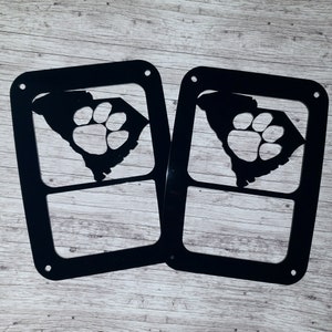 Puede incluir: Dos recortes de metal negro del estado de Carolina del Sur con una huella de pata en el interior. Los recortes est&aacute;n dise&ntilde;ados para ser utilizados como cubiertas de luz.