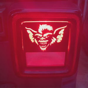 May include: Red vehicle tail light with a glowing, stylized Gremlin face design. The design is in a light color, contrasting against the red light. The tail light is rectangular with rounded corners.