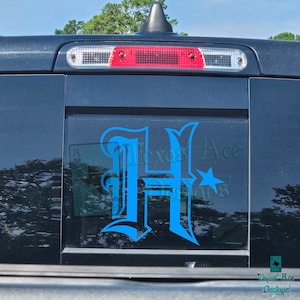 May include: A blue and white decal of a stylized letter H with a star on the back window of a black truck. The decal says "Texas Ace Designs" in smaller text.