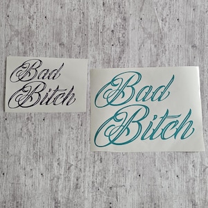 May include: Two stickers with the words "Bad Bitch" in a cursive font. One sticker has black text, and the other has teal text. The stickers are on a light gray and white wood-grain background.