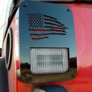 May include: Black metal rear tail light cover with a distressed American flag cutout. The cover is mounted on a red vehicle.