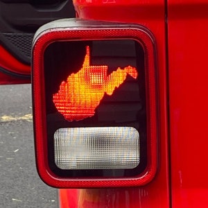 Puede incluir: Un Jeep Wrangler rojo con una pegatina con el contorno del estado de Virginia Occidental en la luz trasera. La pegatina est&aacute; iluminada por la luz trasera, creando un brillo naranja brillante.