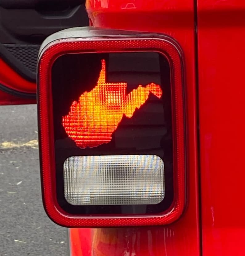 Puede incluir: Un Jeep Wrangler rojo con una pegatina con el contorno del estado de Virginia Occidental en la luz trasera. La pegatina est&aacute; iluminada por la luz trasera, creando un brillo naranja brillante.