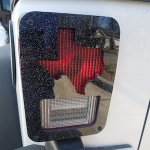Puede incluir: Cubierta de luz trasera negra para Jeep Wrangler con un recorte de contorno del estado de Texas rojo. La cubierta tiene un acabado de brillo negro.