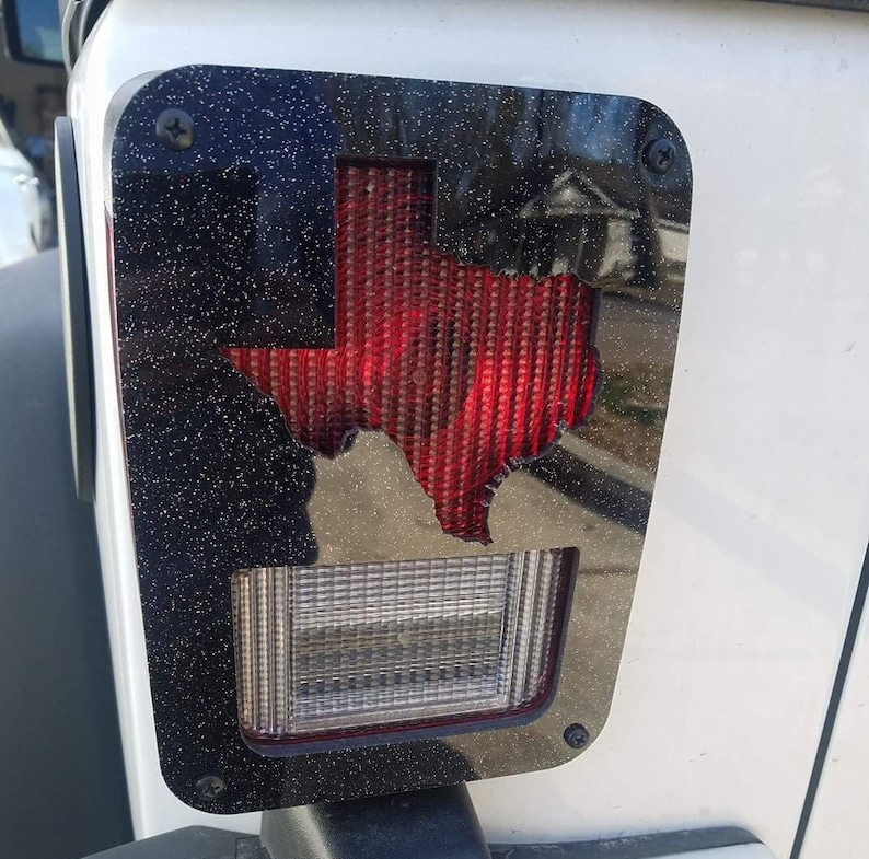 Puede incluir: Cubierta de luz trasera negra para Jeep Wrangler con un recorte de contorno del estado de Texas rojo. La cubierta tiene un acabado de brillo negro.