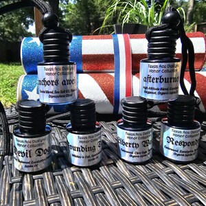 May include: Six small glass bottles with black caps and hanging cords, labeled Texas Ace Designs Honor Collection. The labels feature names like Anchors Aweigh, Afterburner, Devil Dog, Founding, Liberty Walk, and Responder with ingredient lists.