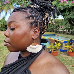 Peut inclure: Une femme porte de grandes boucles d'oreilles à franges crème avec un dessus en bois. Les boucles d'oreilles sont posées sur un haut noir. La femme a les cheveux tressés et est à l'extérieur avec de la verdure en arrière-plan.