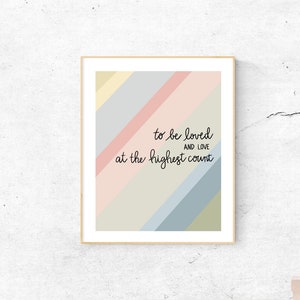 May include: A framed print with a colorful striped background and the text "to be loved and live at the highest count". The print is hanging on a white wall above a white desk with a laptop and a pink vase.