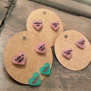 May include: Three sets of heart-shaped earrings with different sayings. The first set is pink and says "Single AF". The second set is pink and says "Can U Not". The third set is green and says "Nah".