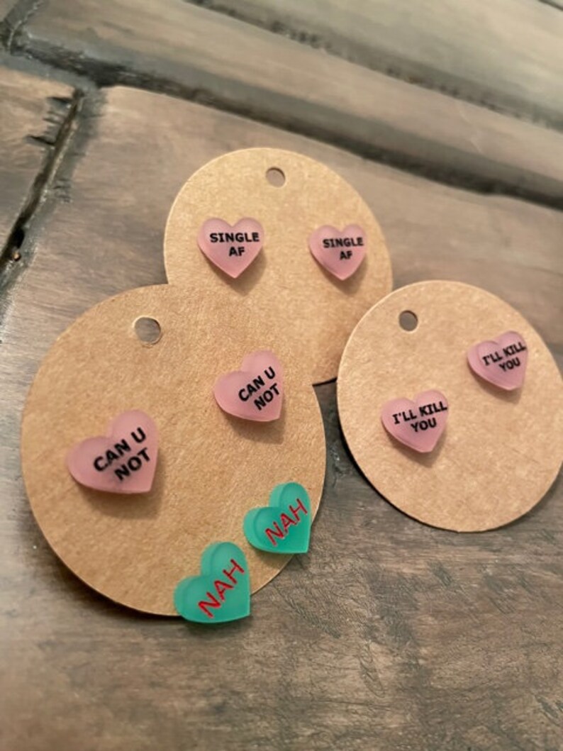 May include: Three sets of heart-shaped earrings with different sayings. The first set is pink and says "Single AF". The second set is pink and says "Can U Not". The third set is green and says "Nah".