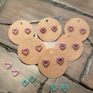 May include: Assortment of pink and green heart-shaped earrings with various phrases printed on them, including "Meh", "Can U Not", "Nah", "Vomit", "I Hate You", "Thank U Next", "Nope", "Be Wine", and "It's Not Me".