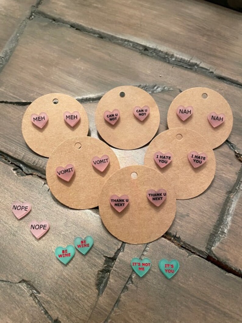 May include: Assortment of pink and green heart-shaped earrings with various phrases printed on them, including "Meh", "Can U Not", "Nah", "Vomit", "I Hate You", "Thank U Next", "Nope", "Be Wine", and "It's Not Me".