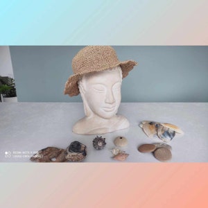 May include: A beige crocheted sun hat sits on a white plaster bust of a person with closed eyes. The bust is on a white surface with seashells and stones.