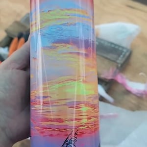 Puede incluir: Un vaso de acero inoxidable con una escena colorida de puesta de sol y playa. La escena presenta un cielo rosa y naranja con nubes, un océano azul y hierba alta.