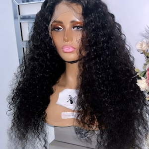 May include: A long, black, curly human hair wig on a mannequin head. The wig is styled in loose curls and falls to the shoulders. The mannequin is wearing a nude-colored wig cap.