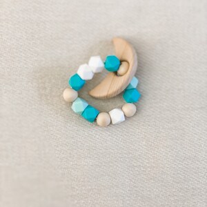 May include: A baby teething toy featuring a crescent-shaped wooden piece and a beaded ring. The beads are a mix of white, teal, and natural wood colors. The toy is designed for infants and toddlers.