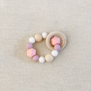 May include: A baby teether with a wooden ring and a beaded design. The beads are in various shapes and colors, including pink, white, and light purple. The teether is designed for infants and toddlers.