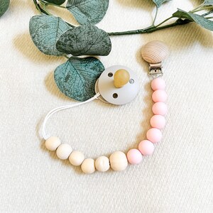 May include: A baby pacifier with a light gray nipple shield and a natural wood-colored nipple. The pacifier is attached to a beaded chain with pink and natural wood beads. A wooden clip secures the chain. Eucalyptus leaves are in the background.