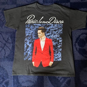 May include: Black t-shirt with a graphic print of a man in a red suit standing in front of a blue floral background. The text "Panic! at the Disco" is printed in white above the graphic.