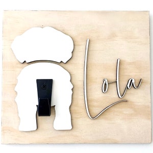 May include: Wooden sign with the name "Lola" in a cursive font. The sign features a white silhouette of a person's head and hair, with a black hook attached. The background is a light wood grain.