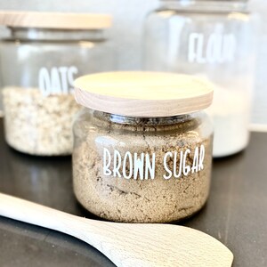 May include: Three glass jars with wooden lids, filled with oats, brown sugar, and flour. The jars are labelled with white text on a black background. The text on the jars reads "Oats", "Brown Sugar", and "Flour". A wooden spoon is in the foreground.
