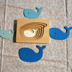 May include: A wooden puzzle with a whale shape. The puzzle has five pieces, each with a different shade of blue. The puzzle is on a white and blue floral fabric background.