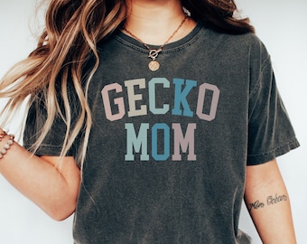 May include: A dark gray t-shirt with the text "Gecko Mom" in colorful letters. The letters are in a rainbow color scheme, with each letter a different color.