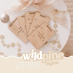 Peut inclure: Un ensemble de cinq étiquettes rectangulaires en bois clair aux coins arrondis, gravées de texte noir incluant DECORATE, BAKE, READ, WRAP et WATCH, accompagnées de petites étoiles. Les étiquettes sont disposées en éventail sur une surface blanche.