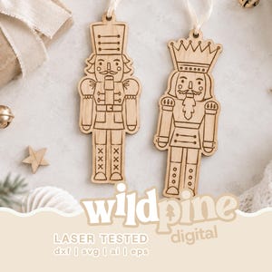 May include: Two light-coloured wooden nutcracker ornaments with laser-engraved details, each featuring a white ribbon loop for hanging. The text "wildpine digital" and "LASER TESTED dxf | svg | ai | eps" is printed at the bottom of the image.