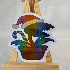 May include: A holographic sticker of a Venus flytrap wearing a Santa hat. The plant is in a brown pot with green leaves and a candy cane. The sticker is displayed on a small wooden easel.