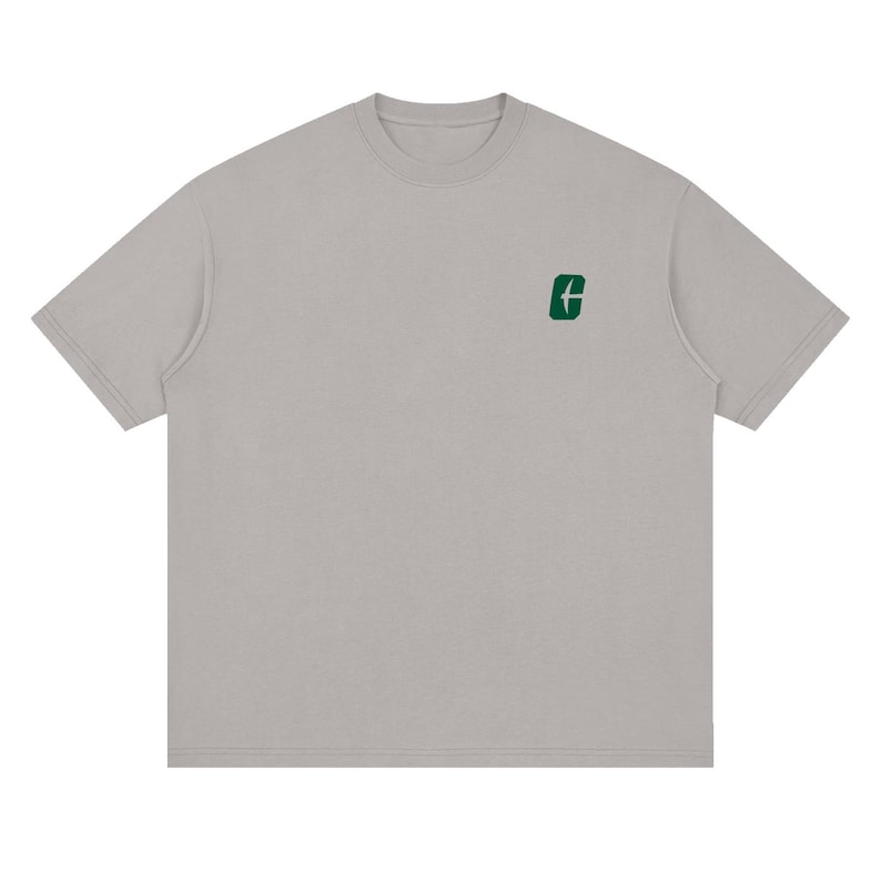 May include: A light gray t-shirt with a crew neck and short sleeves. A small green graphic is on the upper left chest. The shirt has a relaxed fit and a simple, minimalist design.