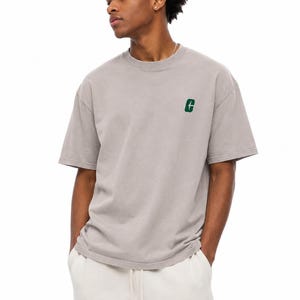 May include: A light gray t-shirt with a small green letter "G" embroidered on the left chest. The shirt has short sleeves and a crew neck. The person is wearing white sweatpants with a drawstring.