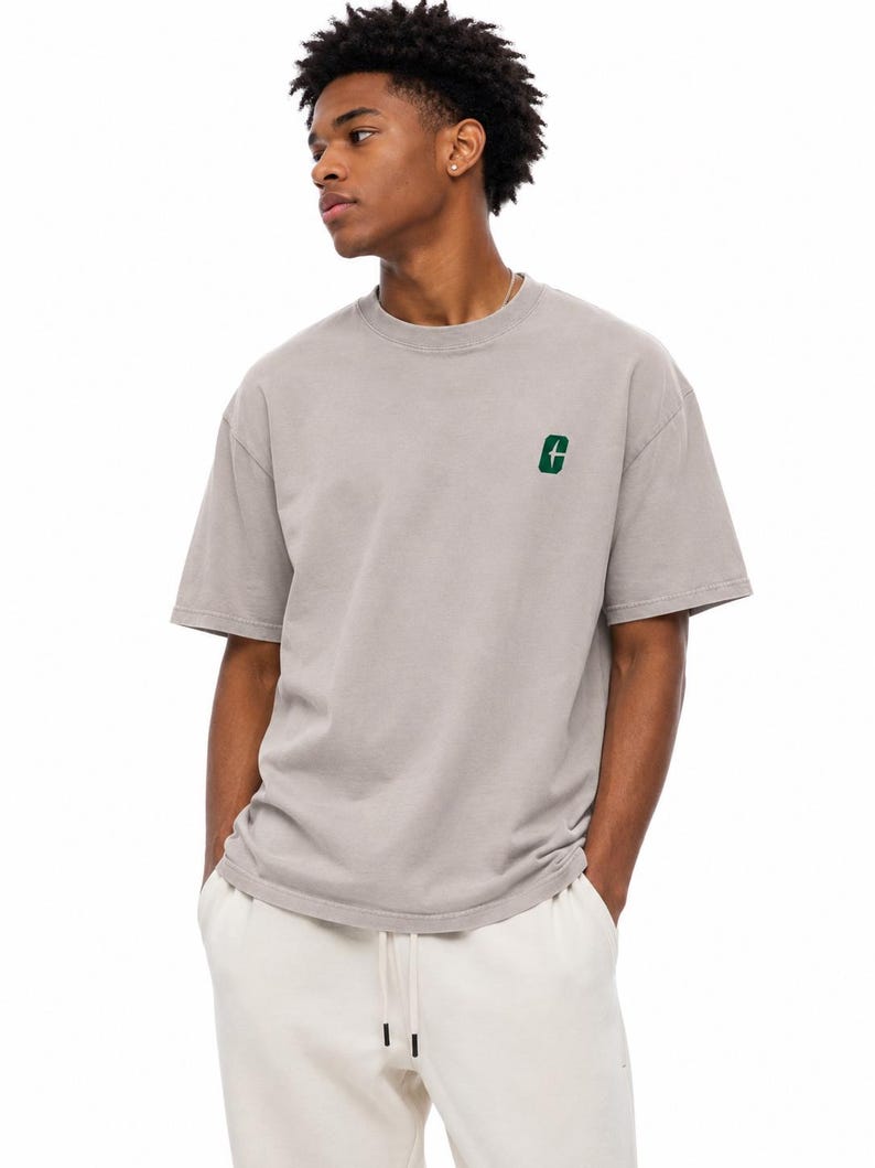 May include: A light gray t-shirt with a small green letter "G" embroidered on the left chest. The shirt has short sleeves and a crew neck. The person is wearing white sweatpants with a drawstring.