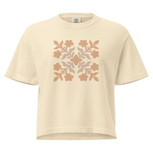 Pode incluir: T-shirt creme com um design floral em tons de castanho e bege. A camisa de manga curta tem gola redonda e um ajuste folgado. O padrão floral é simétrico e centrado na frente da camisa.
