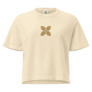 May include: Cream-colored t-shirt with a gold-colored floral design centered on the chest. The short-sleeved shirt has a crew neck and a relaxed fit. The tag on the back of the neck reads "COMFORT COLORS".