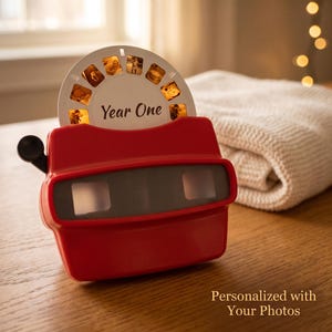 Puede incluir: Un visor estereoscópico de plástico rojo con una palanca lateral negra y un disco fotográfico circular blanco insertado en la parte superior. El disco incluye el texto Year One y contiene pequeñas imágenes. El dispositivo tiene dos lentes rectangulares.