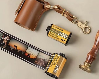 May include: A brown leather film canister holder with a gold-coloured clasp and a matching leather strap. Two yellow Kodacolor 35mm film rolls are next to a strip of developed film with photos. A wooden stamp is also in the image.