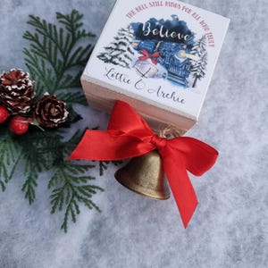 May include: A small wooden box with a printed lid featuring a train, Christmas trees, and the word "Believe". A gold bell with a red ribbon is in front of the box. The text on the box reads "The bell still rings for all who truly" and "Lottie & Archie".