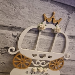 May include: White wooden light switch cover with a gold carriage design, white flowers, and a gold crown. The name "Sophia" is written in black cursive below the carriage.