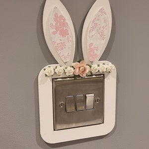 May include: A white light switch cover with bunny ears. The ears are made of white cardboard with a pink floral pattern. The switch cover has a white border and a silver light switch with three buttons.