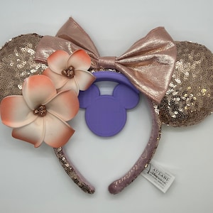 Könnte beinhalten: Roségoldene Minnie Mouse Ohren mit einer rosa und weißen Blumenverzierung und einer lila Schleife. Die Ohren sind aus Pailletten und die Schleife aus Satin.