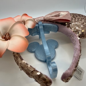 May include: A pair of pink and gold sequined Minnie Mouse ears with a pink bow and a white flower with orange accents. The ears are on a blue plastic stand.