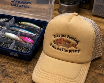 Puede incluir: Gorra trucker color beige con la frase bordada "Take me fishing & tell me I'm pretty" y un gráfico de pez. La gorra tiene la parte trasera de malla y una visera curva. Una caja de aparejos Plano Saltwater y señuelos de pesca en el fondo.