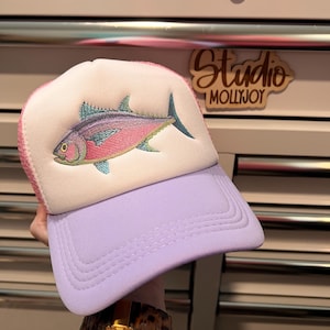 May include: A trucker hat featuring a white front panel with an embroidered pink, purple, and teal fish graphic. The hat has a light purple brim and a pink mesh back. The design includes a structured foam front and a curved visor with stitching.